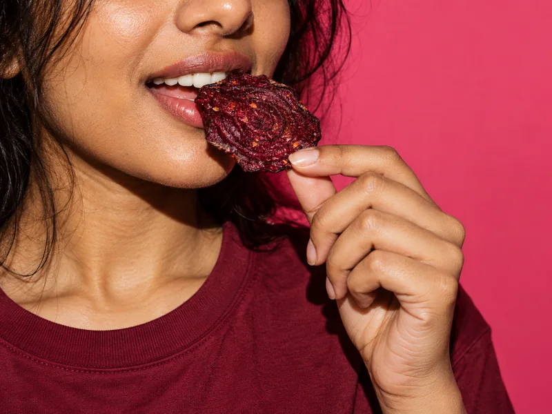 Close-up of a woman about to bite a beetroot crisp against a pink background