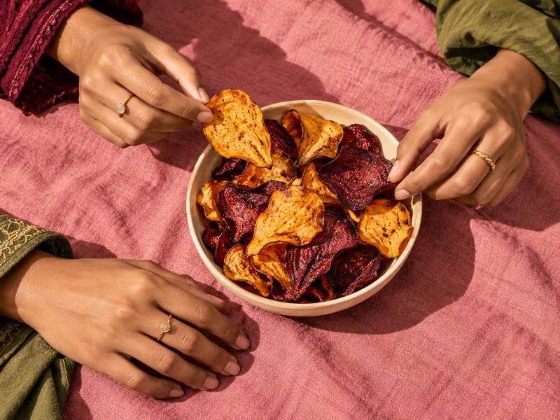Three hands sharing a bowl of beetroot and sweet potato crisps on a pink tablecloth