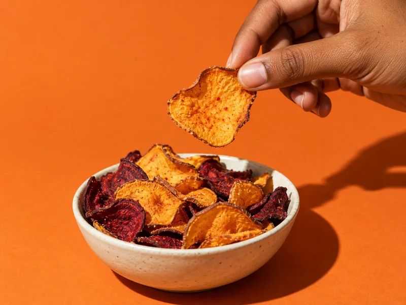 A hand picking a sweet potato crisp from a bowl on an orange background
